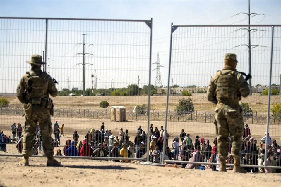 Migrantes esperan junto a una valla fronteriza bajo la vigilancia de la Guardia Nacional de Texas.