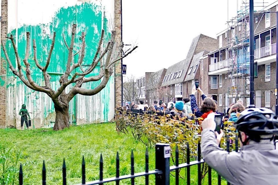 La obra muestra chorros de pintura verde que replican el follaje de un árbol severamente podado y muestra a una figura humana con una manguera de presión.