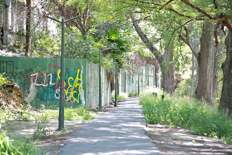 Un andador del parque está descuidado, con la maleza crecida, las luminarias sin farolas y las paredes pintadas con grafiti.