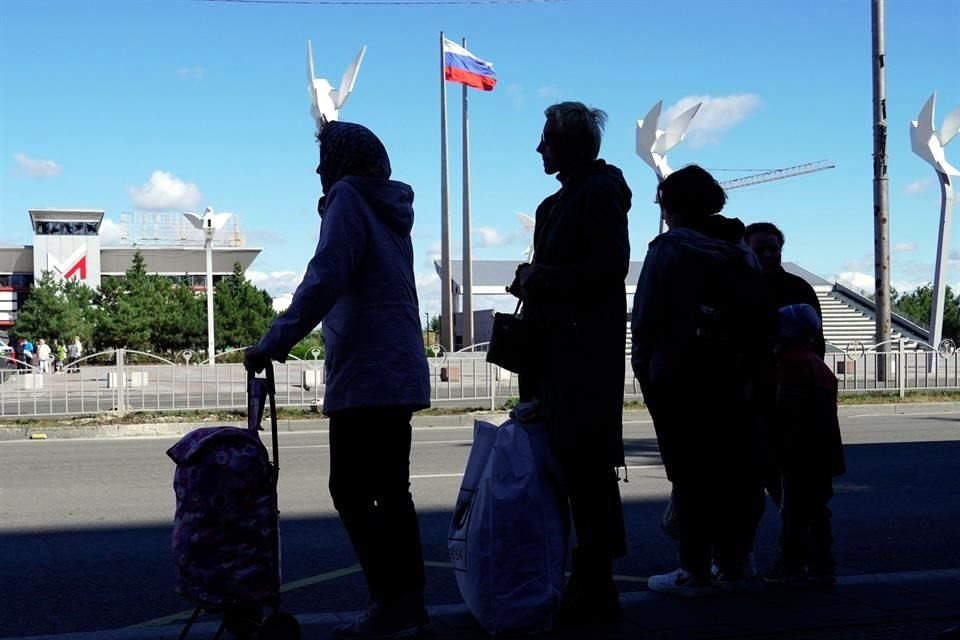 Pasajeros en Mariúpol, con una bandera rusa al fondo, esperan un autobús.