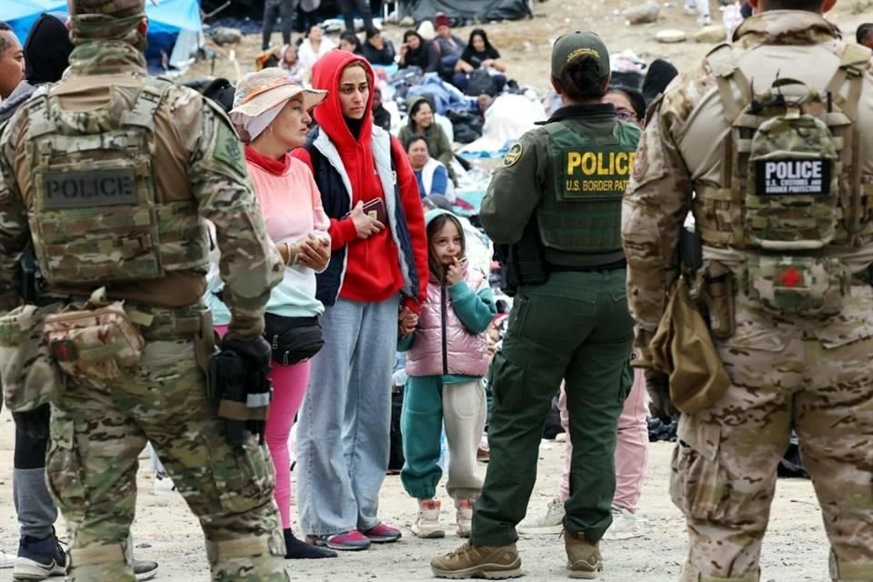 Agentes y oficiales federales  vigilan a los migrantes que hacen fila para ser transportados desde un campamento improvisado en la frontera de EU y México.